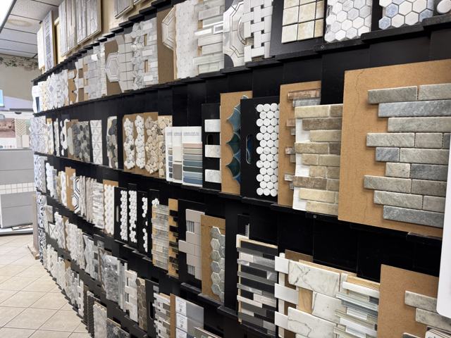 A long wall of mosaic and accent tile samples: marble hexagons, basketweave, penny rounds, pebble stone, glass linear, and blue fan tile